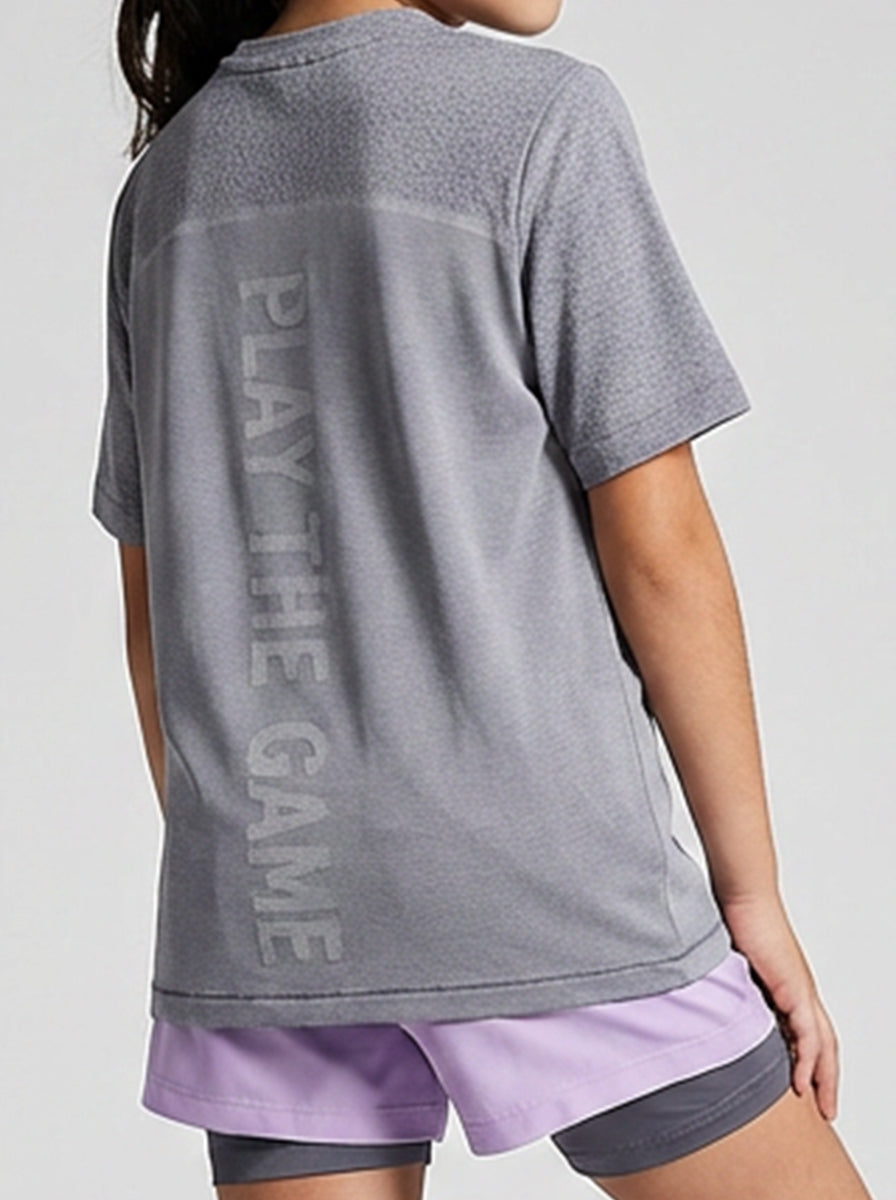 A girl wearing a gray t-shirt with 'Play the Game' text on the back and purple shorts.